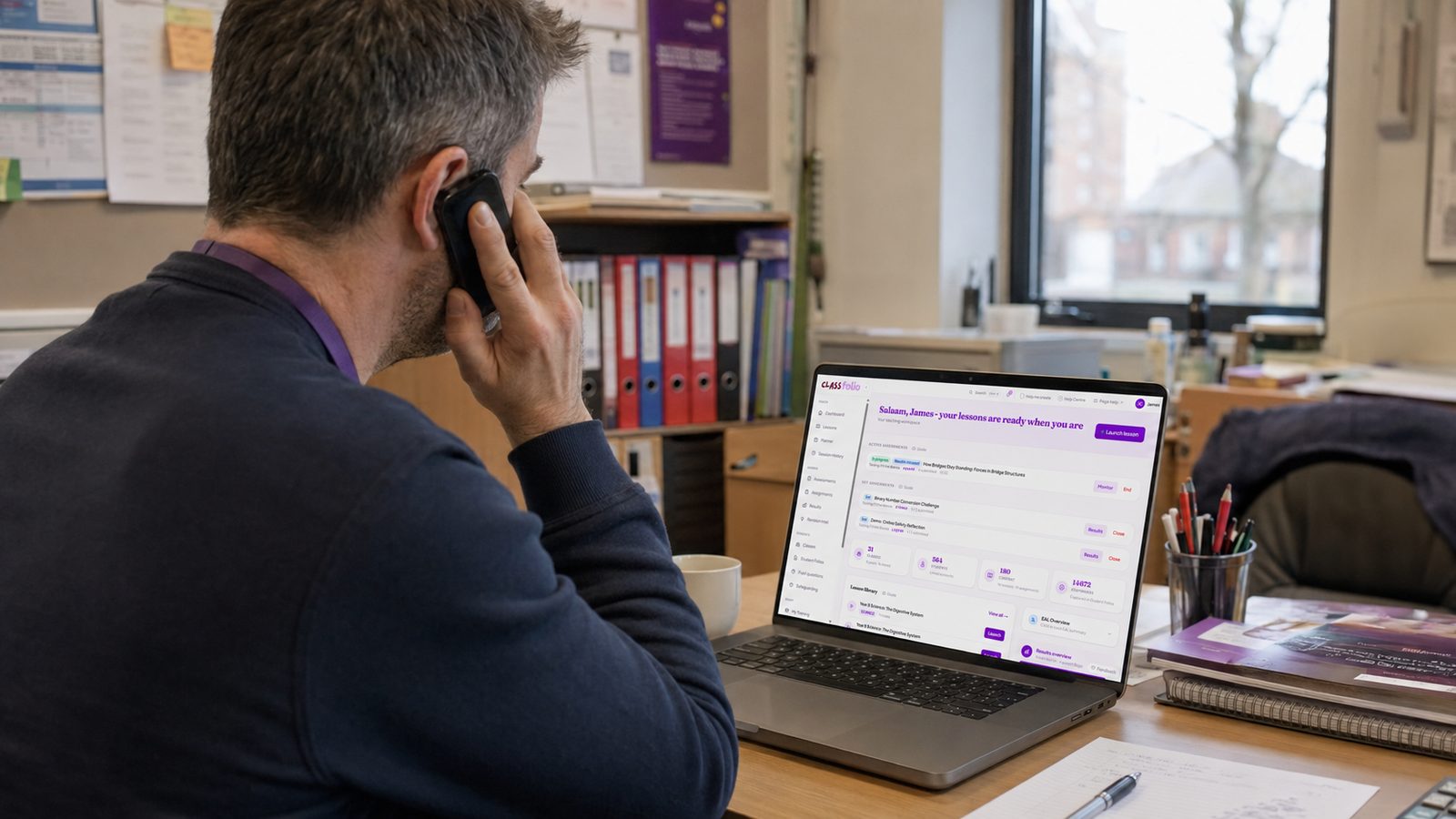 A teacher sitting at a desk in a school office, holding a phone to his ear while looking at a laptop showing his Classfolio dashboard.